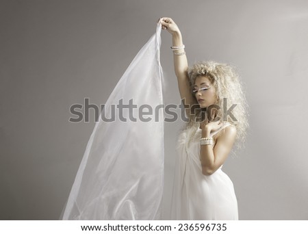 Blonde woman with white fantasy makeup and hairstyle holding up white material blowing in the breeze