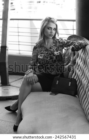 Sexy blonde woman seated on couch in urban setting with black handbag next to her in black and white.