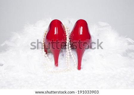A pair of sexy, red patent leather pumps sit atop a white fur surface with a white background; pearls and a white boa surround the shoes