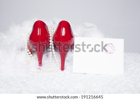 A pair of sexy, red patent leather pumps sit atop a white fur surface with a white background; pearls and a white boa surround the shoes; a love note with a lipstick kiss sit to the side