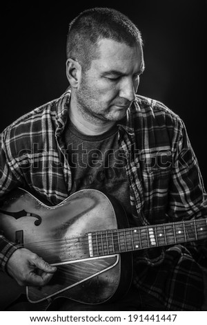 A man wearing a plaid flannel shirt plays an old acoustic guitar; black and white photo