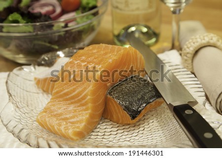 Two pieces of fresh salmon sit atop a plate ready to be cooked.  A sharp knife rests on the side of the plate.