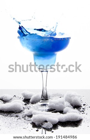 A margarita glass is filled with a blue frozen margarita; white sugar coats the rim; slushy snow sits around the base of the glass and is reflected off the surface;