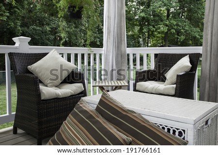 Two resin wicker chairs sit on a covered deck; they have cream colored cushions and pillows; canvas curtains hang in the background; a white wicker chest serves as a coffee table