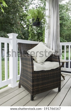 A resin wicker chair sits on a covered deck with cream colored cushion and pillows; curtains hang behind; plants hang from above