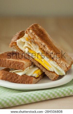 A fried egg sandwich on whole grain toast sits on a white plate atop a green and white checked napkin and a wooden cutting board.