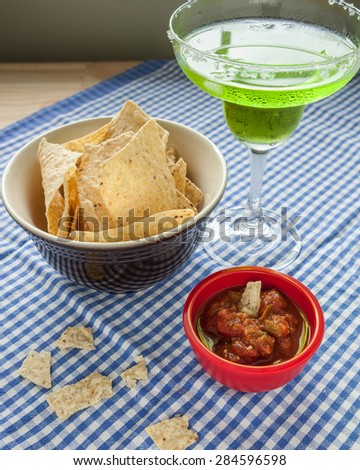 A bowl of corn tortilla chips sit beside a red bowl of salsa on a blue and white checkered tablecloth.  Pieces of tortilla are broken in the foreground.  A bright green margarita is also pictured.