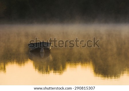 An aluminum row boat moored offshore on a lake at sunrise with fog rising off the lake and trees reflected on the waters surface.