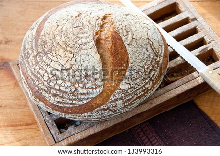 A large whole grain loaf of rustic country bread rests on an old, much used bread board. Crumbs of previous loaves are visible through the slats of the bread board.