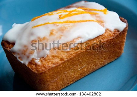 A lemon frosted almond cake sits on a blue plate. The cake is topped with a thin strip of lemon rind.