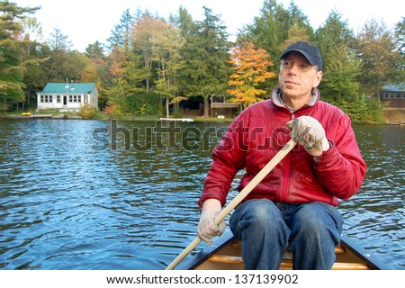 A man paddles a canoe on a lake in Vermont during fall foliage season. Summer cabins line the shore in the background.