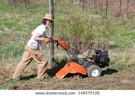 A woman in work clothes pushes a rototiller in a garden. She is wearing hearing protection and heavy gloves.