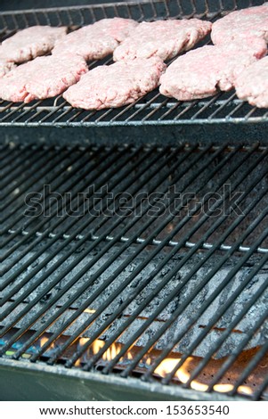 Fresh hamburger patties are laid out on a backyard grill with flames.
