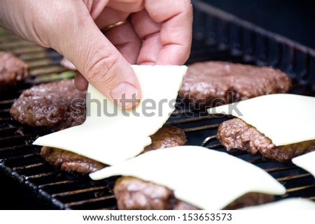 A hand lays out slices of white cheese on freshly grilled hamburgers.