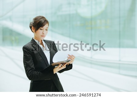Attractive female Asian business executive in a stylish jacket standing holding a clip board in front of a cool toned reflection on an urban building facade, with copyspace