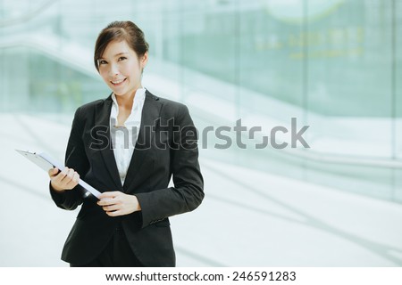 Attractive female Asian business executive in a stylish jacket standing holding a clip board in front of a cool toned reflection on an urban building facade, with copyspace