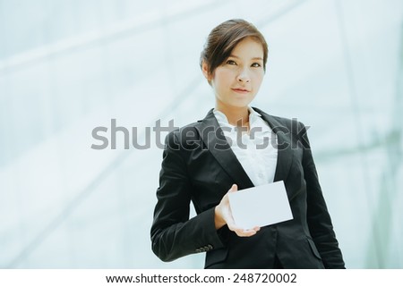 Attractive female Asian business executive in a stylish jacket standing holding a white card in front of a cool toned reflection on an urban building facade, with copyspace