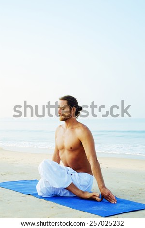 Long hair athletic man with no shirt doing yoga on blue mat at the beach Gomukhasana - Cow face pose with hands down