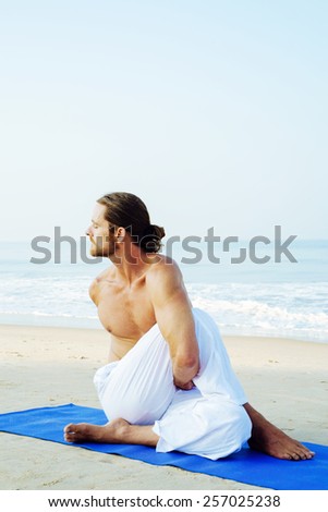 Long hair athletic man with no shirt doing yoga on blue mat at the beach Ardha Matsyendrasana - Seated twist posture