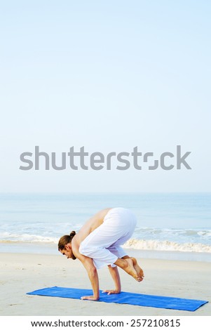 BAKASANA - CROW POSE Long hair athletic man with no shirt doing yoga on blue mat at the beach