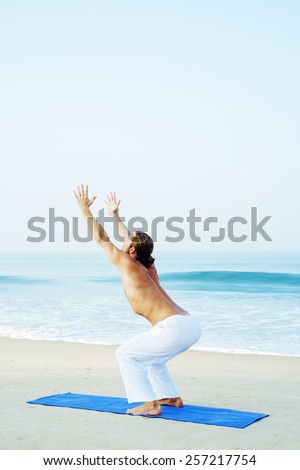 UTKATASANA - CHAIR POSE Long hair athletic man with no shirt doing yoga on blue mat at the beach