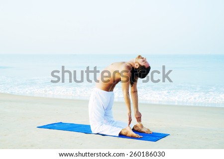 Long hair athletic man with no shirt doing yoga on blue mat at the beach