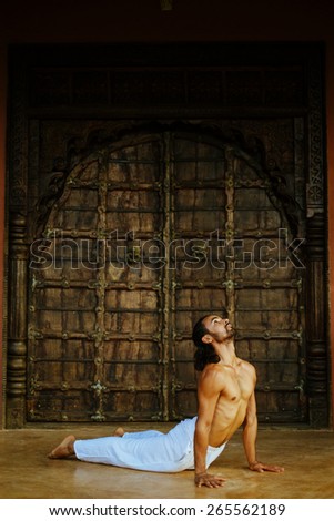 Indian ethnicity young man wit a strong body showing cobra yoga pose (Bhujangasana) in front of old oriental style door.