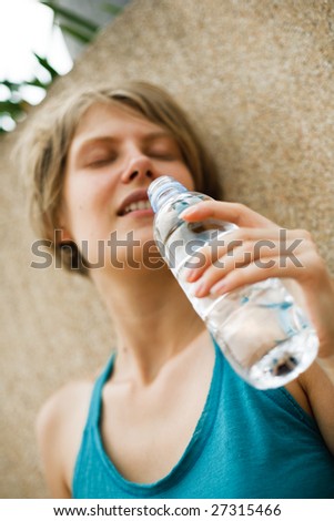 young blond woman with closed eyes drinking water from plastic bottle standing near the wall (blurred face)