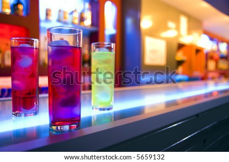 Three cocktail drinks on a bar, blurry color background