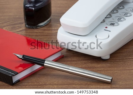 A white phone, an inkwell, a red notebook and a fountain pen on wooden texture imitating a desk.