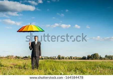 Businessman standing in a green field with multicolored umbrella.