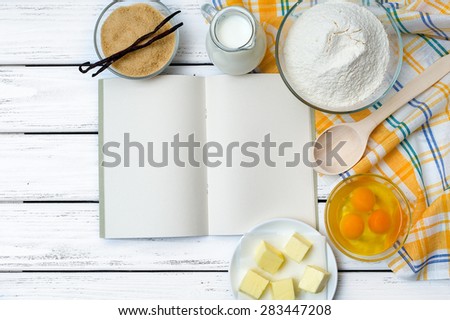 Baking cake in rural kitchen - dough recipe ingredients (eggs, flour, milk, butter, sugar) with recipe book, wooden spoon and kitchen towel on white wooden table from above.