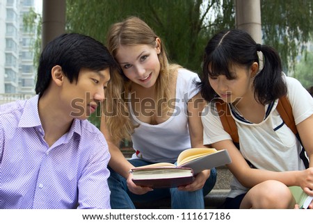 Woman holding books and smiling. Group of young teen students looking at book. People working together in summer park