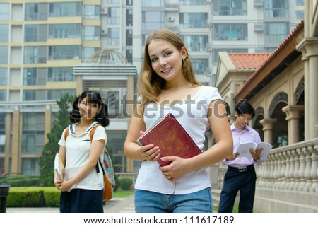Young girl holding book and smiling. Group of students one man and two woman  in park. American and Asian teenagers studying outside of university campus