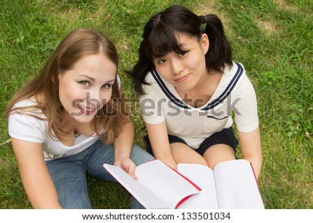Happy university women student outdoor sitting on grass. Asian and Caucasian girl reading book in the park