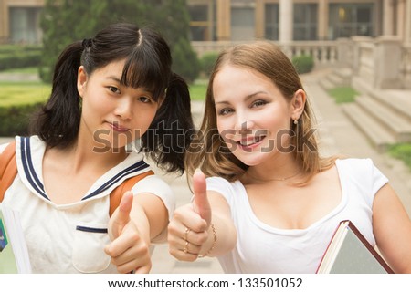 Students in park smiling and showing happy thumbs up success sign. Asian Caucasian women university student holding books outside