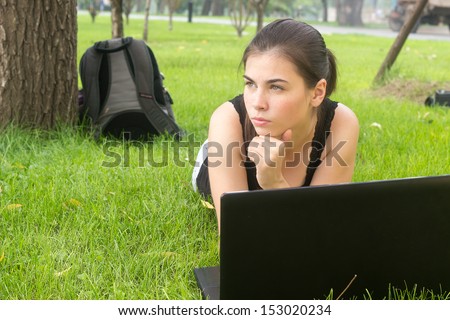 College student lying down on grass and working on laptop at campus park, think and look serious by doing homework