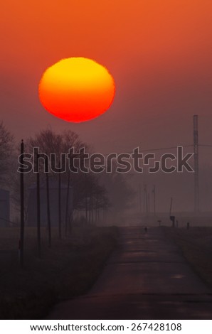 The sun rises above the trees and grain bins along a rural road, on a foggy morning
