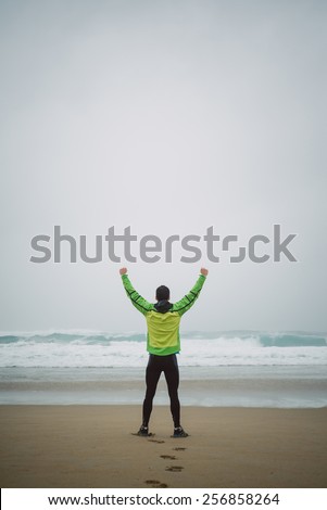 Runner man on the shore of the beach with hands up in victory symbol in a rainy day