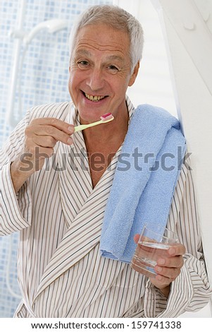 Senior man on his bathroom , towel on the shoulder brushing his teeth with a glass of water on this left hand