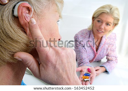 Senior Woman Inserting A Hearing Aid In Her Hear. Focus On The Colored Hearing Aid In Specialist\'S Hand