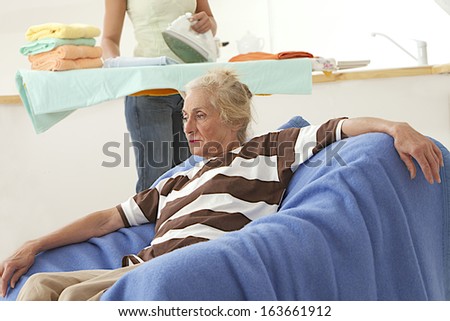 Closeup of a thoughtful elderly woman with home help in background ironing