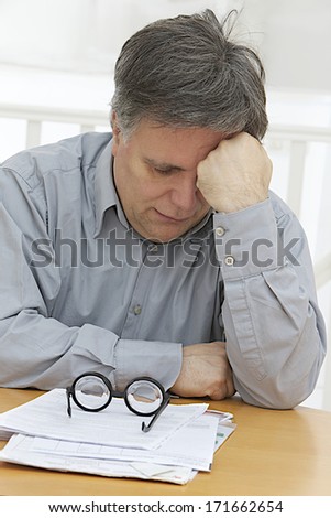 Man at desk holding his head and worrying, his glasses down the desk