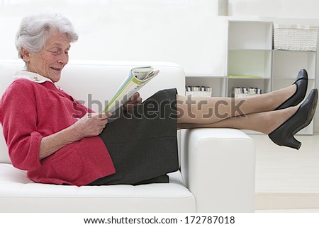 Smiling elderly woman reading on a sofa relaxing with her bare feet over the arm couch