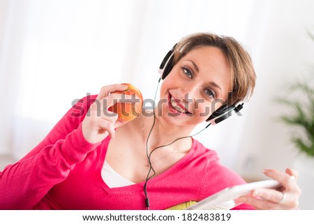 pretty young women on a sofa, listening music on her tablet computer and eating an apple