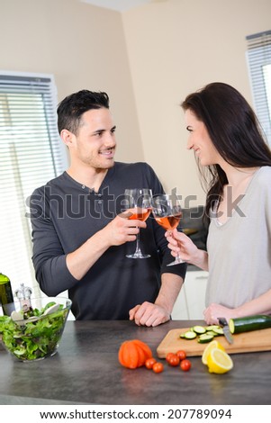 happy beautiful young couple together in kitchen cheering with a glass of wine while preparing salad with organic vegetables for lunch summer party