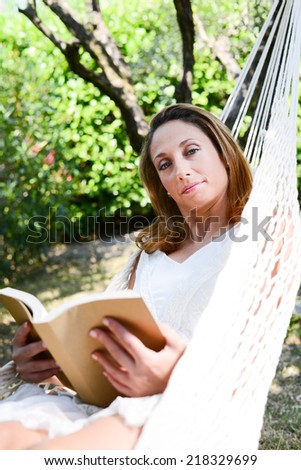 cheerful young woman relaxed reading a book in a hammock in a peaceful garden during summer holiday
