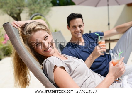 beautiful young couple having drink in deck chairs by the pool in holiday resort