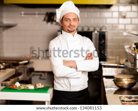 Confident young male chef posing with arms crossed, kitchen background.