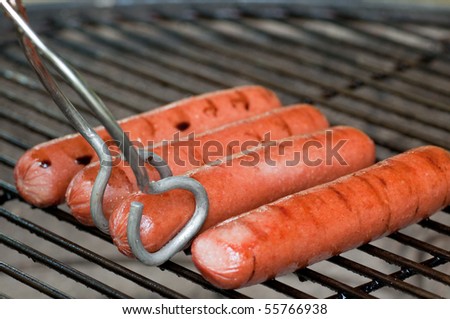 Four hot dogs on a charcoal grill with tongs about to turn one